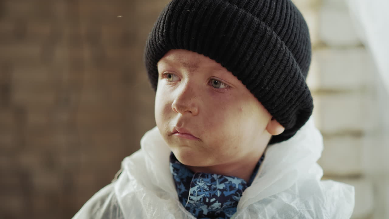Close up of young boy with dirty face wearing black knitted hat and white protective jacket, staring ahead with serious focused expression against blurred background