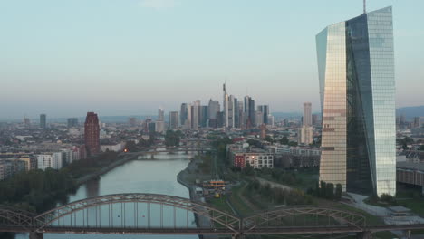 European-Central-Bank-in-early-morning-sunlight-reflection-and-Frankfurt-am-Main-Skyscrapers-in-background,-Aerial-high-angle-forward,-slow