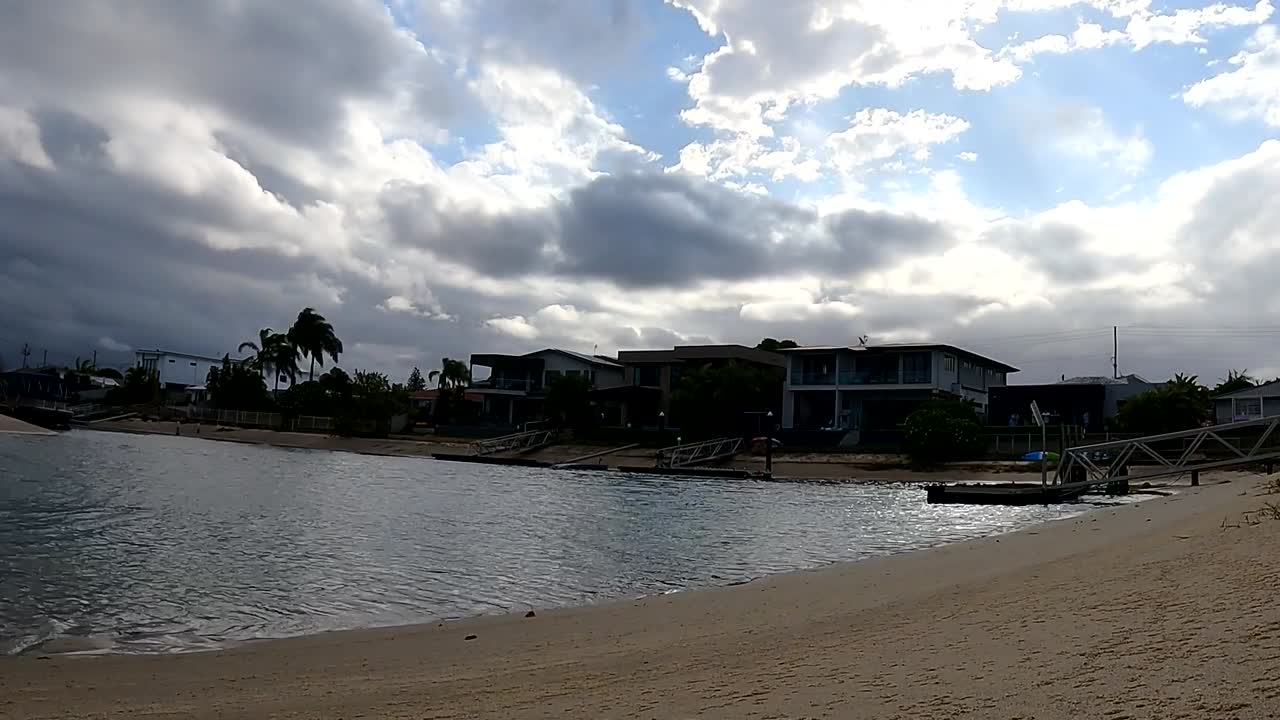 A serene view of clouds moving over waterfront homes and a sandy beach, reflecting light on the water.