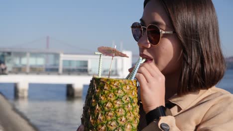 close-up asian woman drinks a cocktail out of a pineapple on a pier in portugal