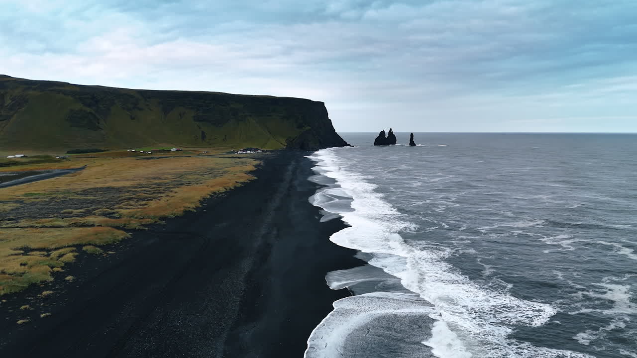 Contrasting white waves splash on the black sandy beach of volcanic origin. Plain mount and few separate rocks sticking out of water at backdrop. Aerial perspective on Iceland shore.
