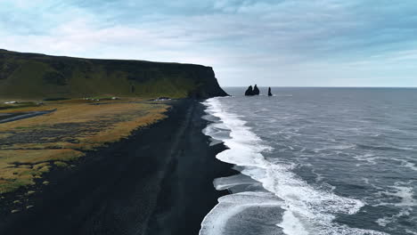 contrasting white waves splash on the black sandy beach of volcanic origin. plain mount and few separate rocks sticking out of water at backdrop. aerial perspective on iceland shore.