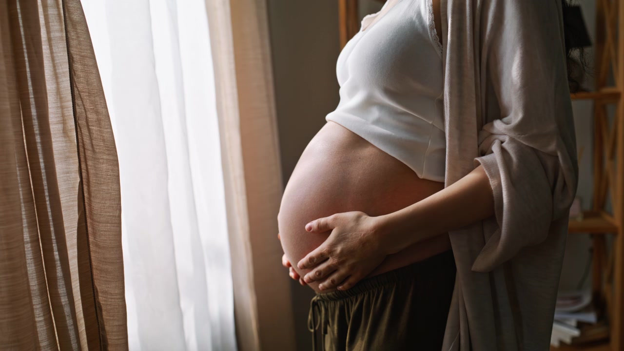 Donna incinta in piedi alla finestra di casa, che si tiene la pancia. Madre serena che si gode la luce.