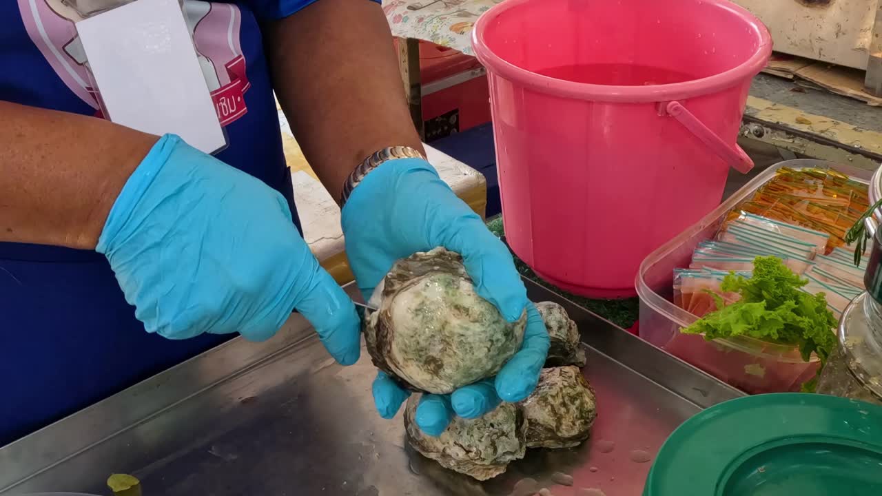 A vendor skillfully handles artichokes, using gloves and a knife, with a pink bucket and fresh greens nearby.