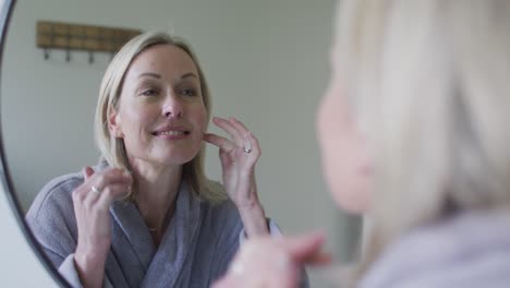 happy senior caucasian woman in bathroom, looking to mirror, touching face