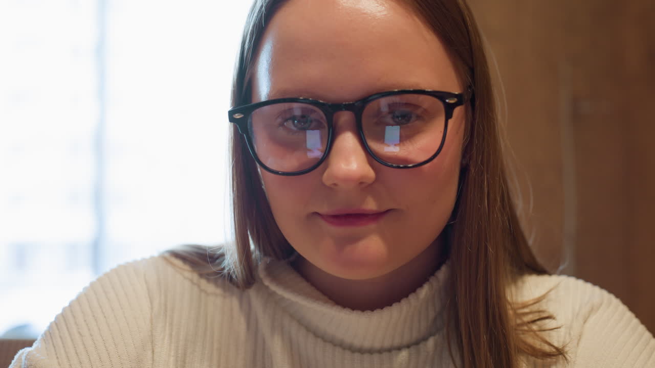 Lady in white ribbed sweater wearing black glasses looks up with a gentle smile while seated indoors near bright window, phone screen reflection visible on lenses in warm wooden interior