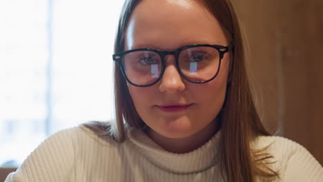 lady in white ribbed sweater wearing black glasses looks up with a gentle smile while seated indoors near bright window, phone screen reflection visible on lenses in warm wooden interior