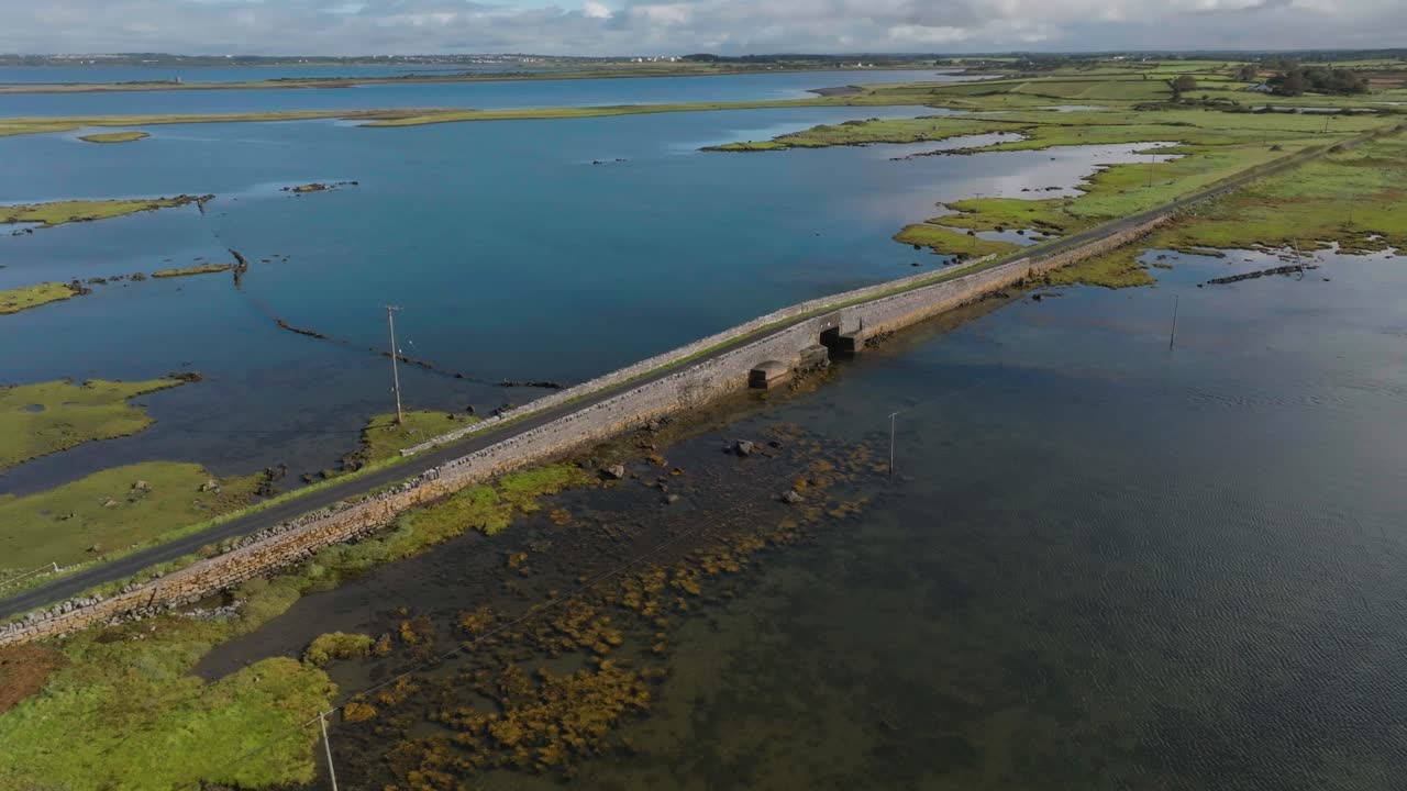 A highangle drone shot showing a long stone bridge traversing a calm coastal lagoon and marshland under a cloudy sky. Ideal for travel and environmental documentaries