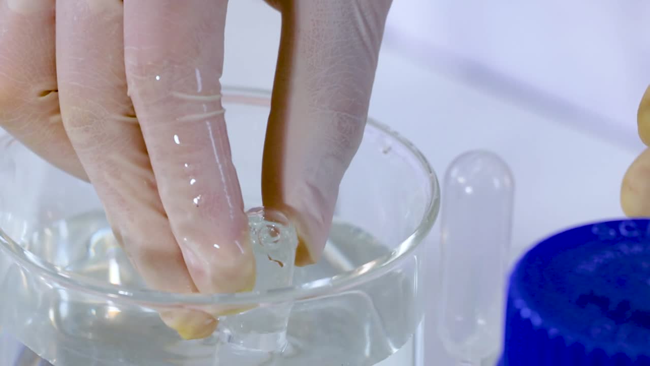 Close-up of gloved hands manipulating glassware in a liquid-filled beaker with blue-capped bottles nearby.