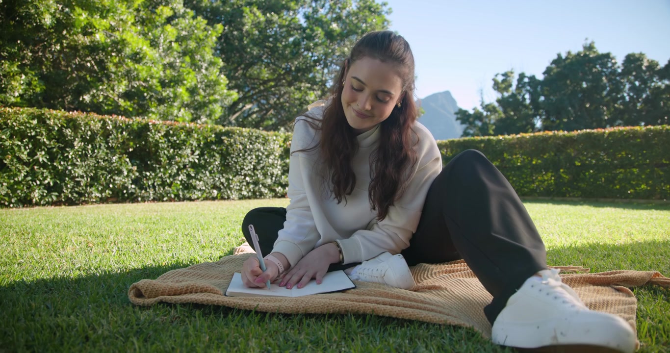 Woman settling on tan blanket, leaning forward, studying with pen in notebook on lawn