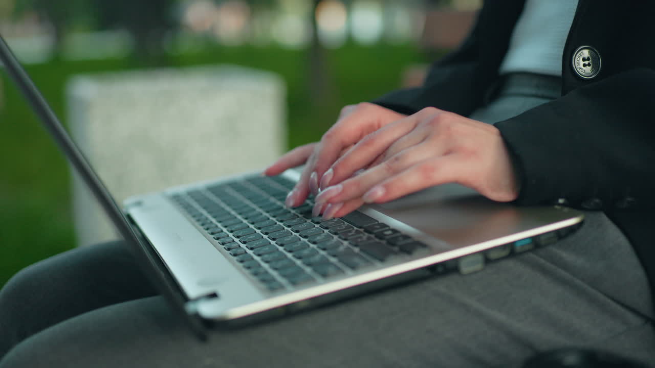 Close up of lady with polished nails typing on laptop in outdoor park setting wearing formal blazer seated on bench with blurred green trees in background