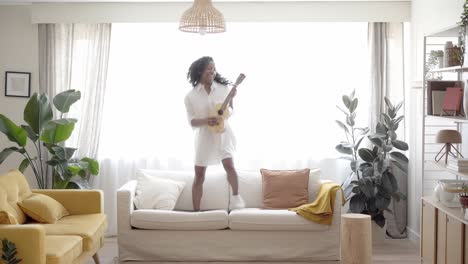 motivated woman playing ukelele standing and jumping on the sofa living room. beautiful latin girl comfortable at home enjoying cheerful music