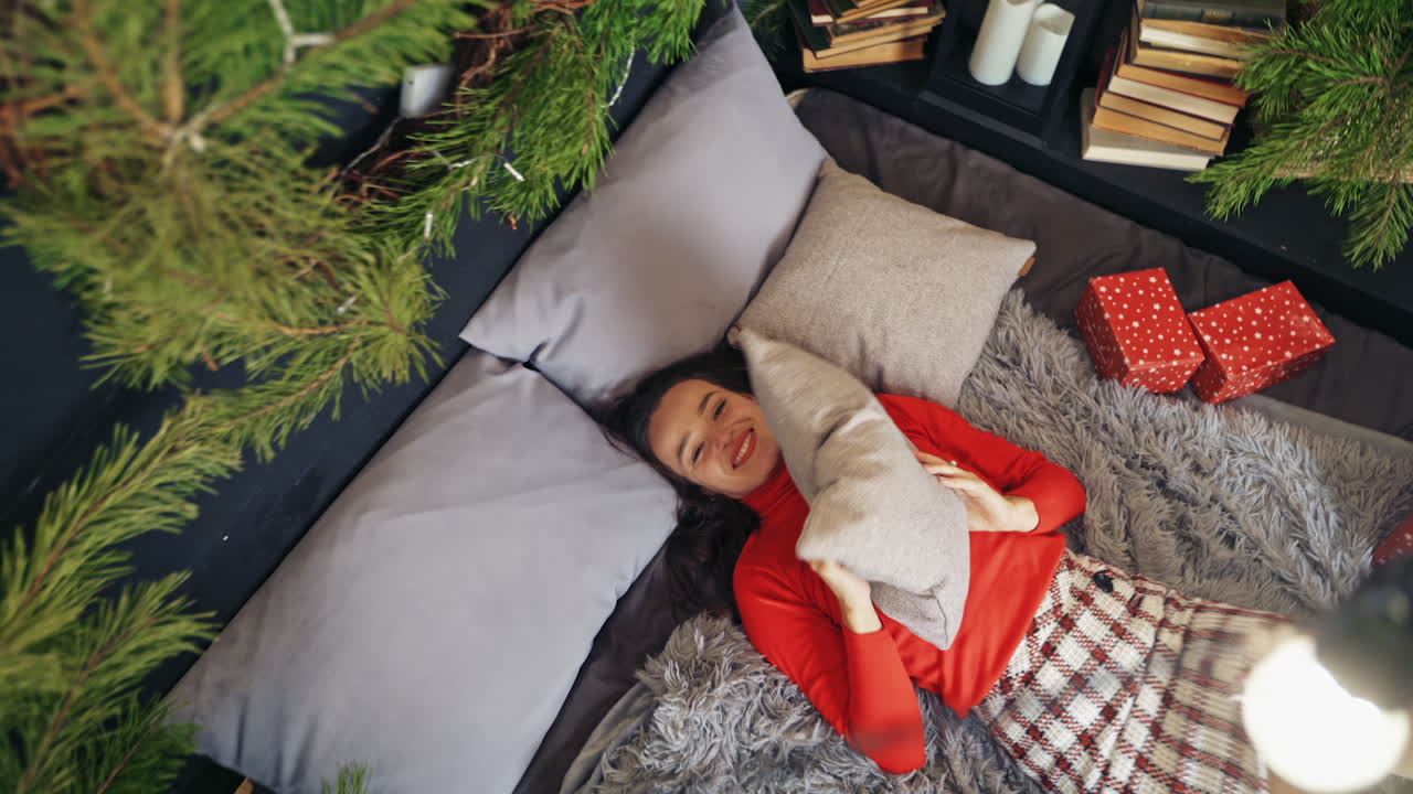 Cute woman throws pillow to the air laying on bed. Room decorated before Christmas. Video from the top.