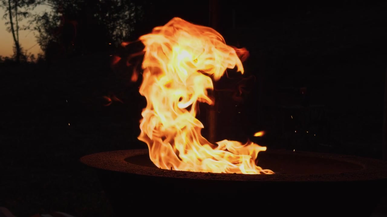 Bright orange flames burning in an outdoor fireplace during the late evening. Close view of glowing fire creating warmth and light against a dark background.