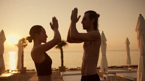 A-brunette-girl-in-a-black-top-and-a-brunette-guy-are-meditating-on-a-Sunny-beach-in-the-morning-and-holding-their-folded-hands-in-front-of-their-chests.-Namaste-position-during-meditation-against-the-backdrop-of-a-golden-sunrise