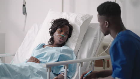 african-american woman resting in hospital bed after surgery talking to young male nurse. portrait of african-american nurse assisting ill female patient lying in bed