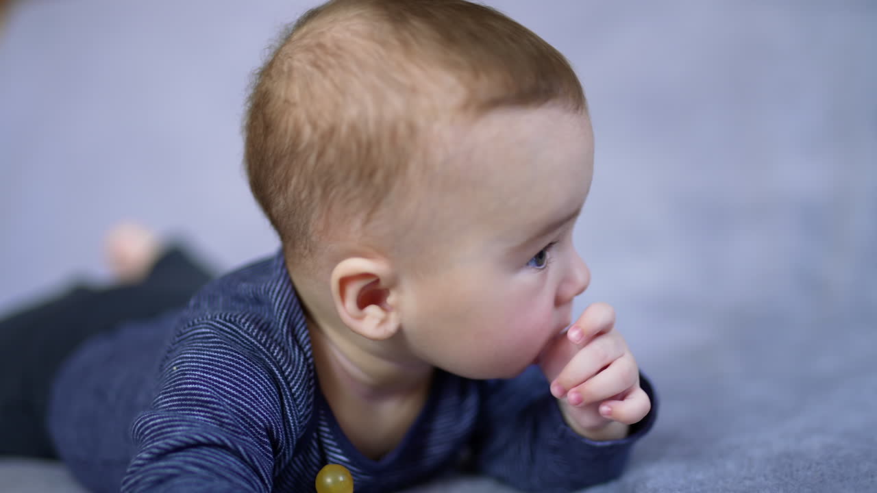 Sweet Caucasian toddler boy lying on belly with a pacifier in front of him. Cute kid looks aside holding his thumb in mouth.