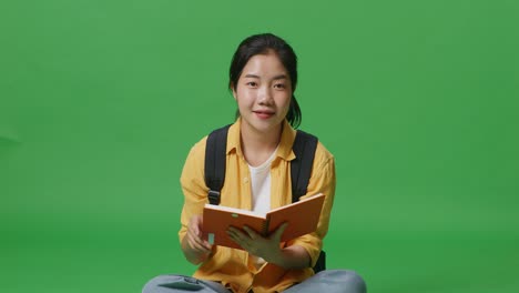 asian woman student with a backpack reading a book then smiling to camera while sitting in the green screen background studio