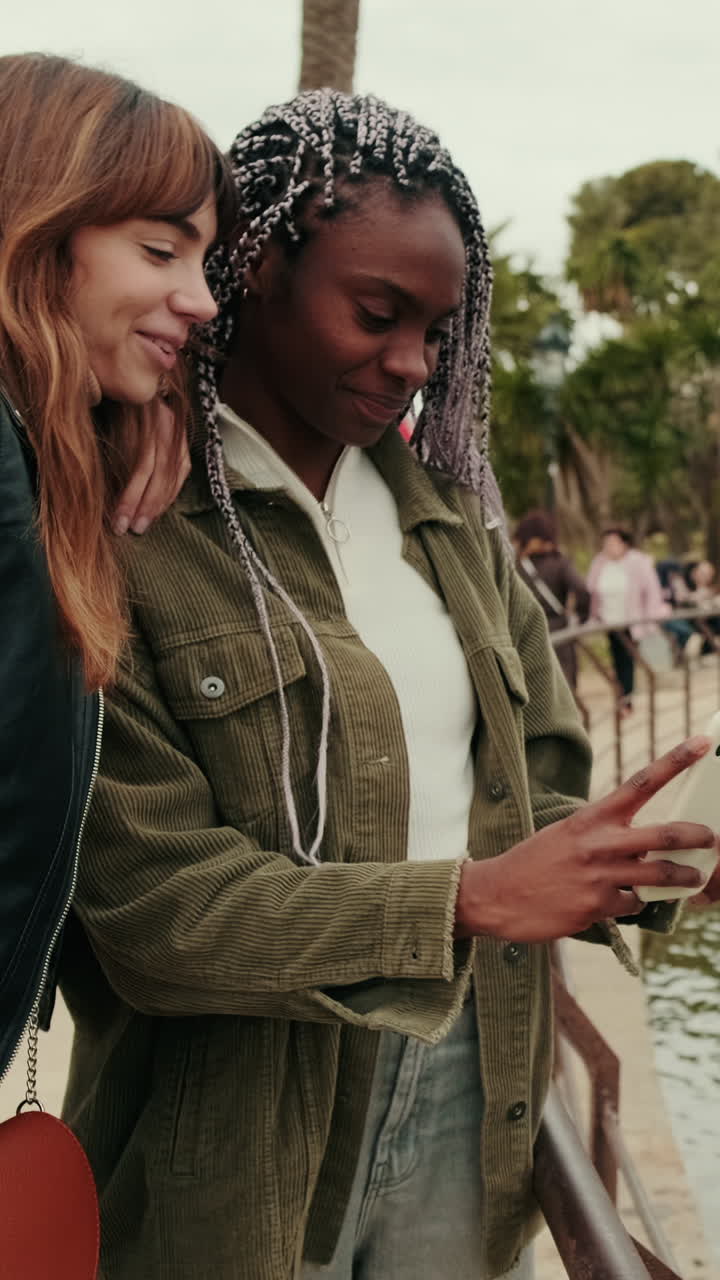 Zwei junge Frauen lächeln und teilen Inhalte auf einem Smartphone in einem Park