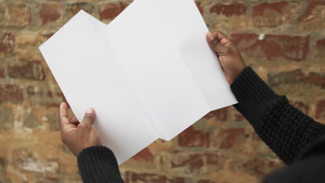 video of hand of african american man holding white blank page, copy space over brick wall