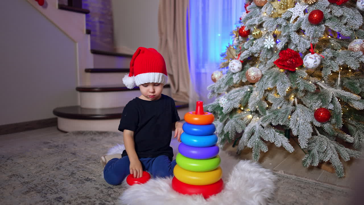 Lovely baby assembles a pyramid and then pushes it fall. Toddler in Santa cap playing near the Christmas tree.