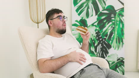 boy touching his belly fat and eating a sandwich sitting on sofa