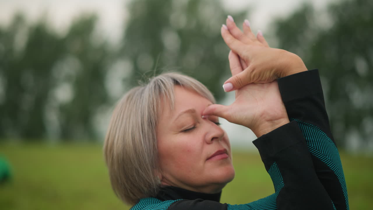 de mediana edad con las manos retorcidas y tocando su frente, ojos cerrados, practicando yoga al aire libre en un campo de hierba brumosa, rodeada de árboles, material verde y amarillo visible en un fondo borroso