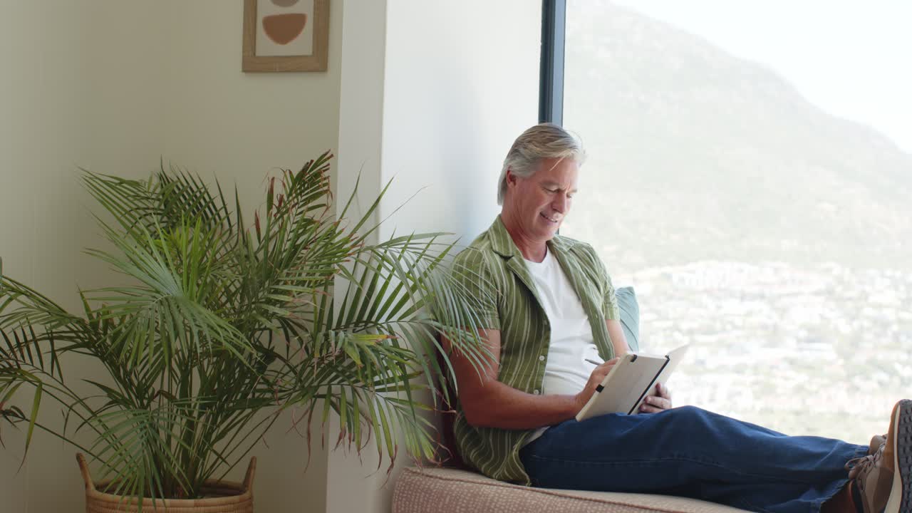 Senior man sitting on window bench, gazing at hillside view, using stylus on tablet, seeking rest