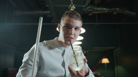 close-up of guy wearing white shirt holding cue stick and glass of lemon drink with black straw. he raises the glass and takes a sip, with overhead lighting in a stylish pool hall setting