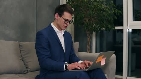 Caucasian-businessman-wearing-formal-blue-suit-and-stylish-glasses-sitting-on-the-sofa-typing-on-his-laptop-on-knees-at-the-hotel-room.-Loft-interior-room
