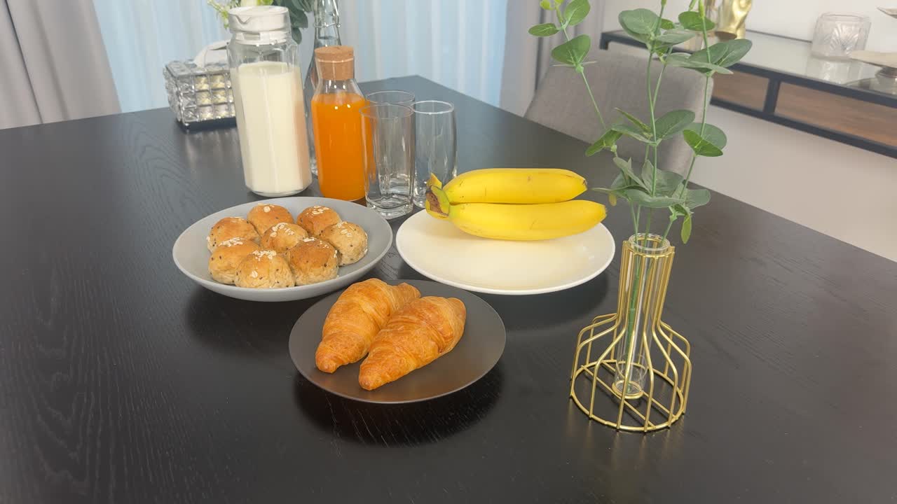 A beautifully arranged breakfast table with bananas, croissants, milk, and orange juice in a well-lit dining room