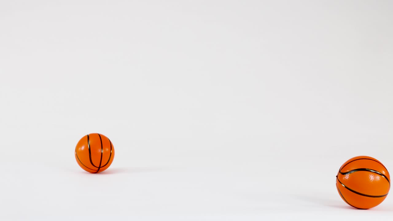 Two miniature orange basketballs bounce and roll across a clean white studio floor surface
