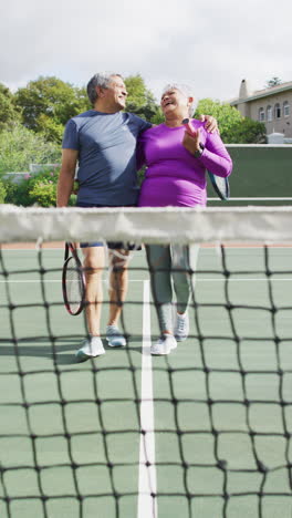 video vertical de una feliz pareja biracial de mayores abrazándose en la cancha de tenis