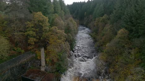 Drohnenaufnahmen-Drehen-Einen-Schnell-Fließenden-Fluss-Und-Einen-Wasserfall-Hinab,-Umgeben-Von-Alten-Gebäuden-Und-Einem-Wald-Aus-Laub--Und-Nadelbäumen