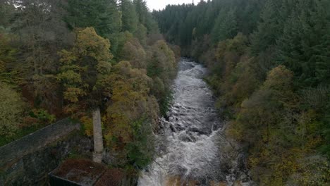 drone footage reverses down a fast flowing river and waterfall surrounded by old buildings and a forest of broadleaf and coniferous trees