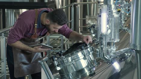 young male brewer wearing a leather apron supervise the process of beer fermentation at a modern brewery factory