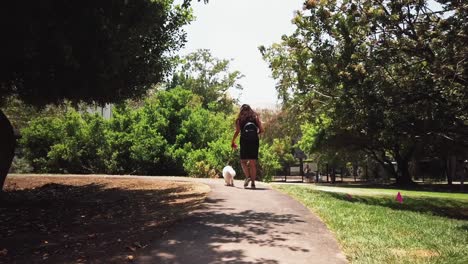 female walking into frame with small white dog in park, on a path, with surrounding trees, los angeles