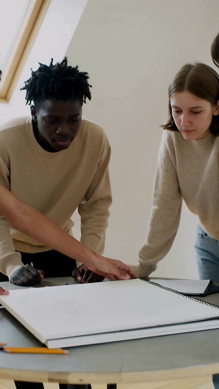 Video verticale: Colleghi chinati che esaminano uno sketchbook mentre la mano di una terza persona apre le pagine in studio