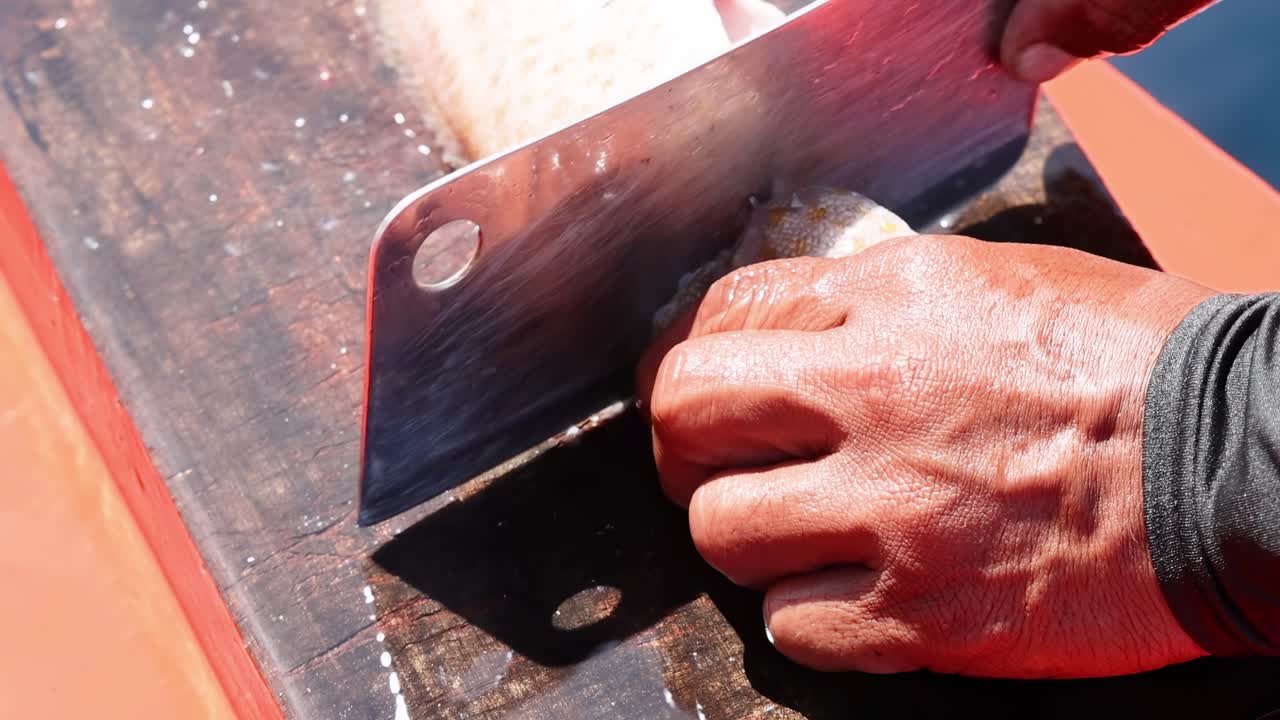 A person skillfully cuts a fish head using a cleaver on a wooden board in bright sunlight.
