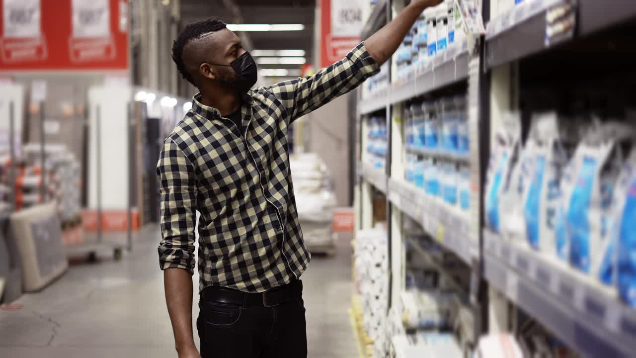 A man in mask walk along the shelves in the store and choose a product