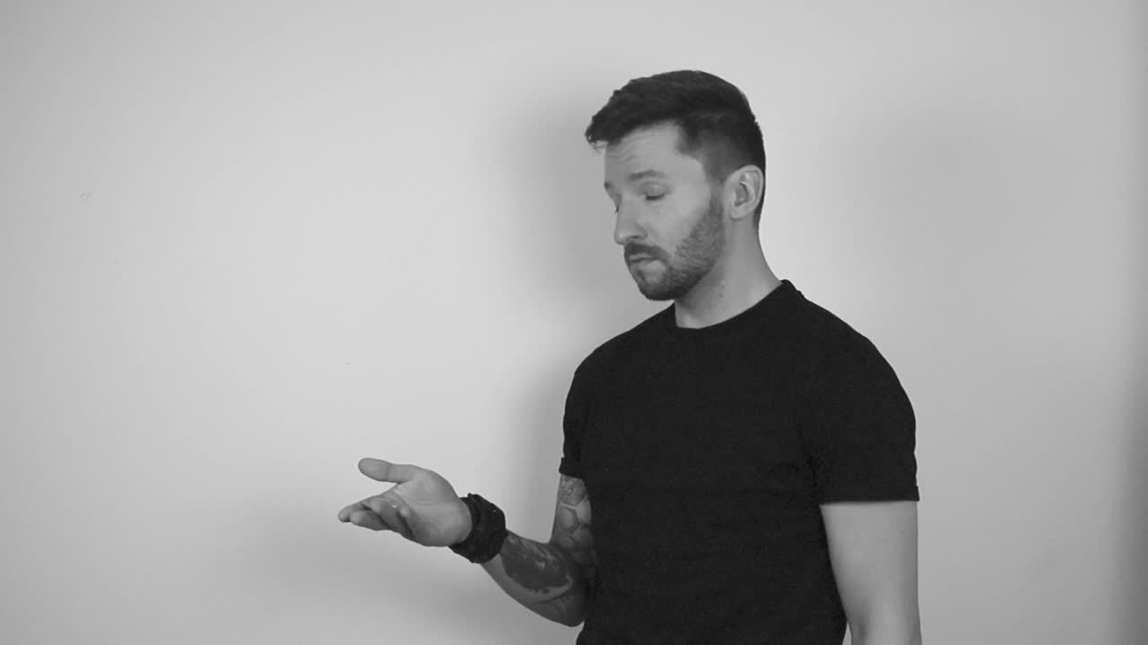 Grayscale footage of a tattooed man spinning a Bitcoin coin in slow motion by throwing it into the air and catching it. After catching it the dude shows the coin to the camera while watching directly.