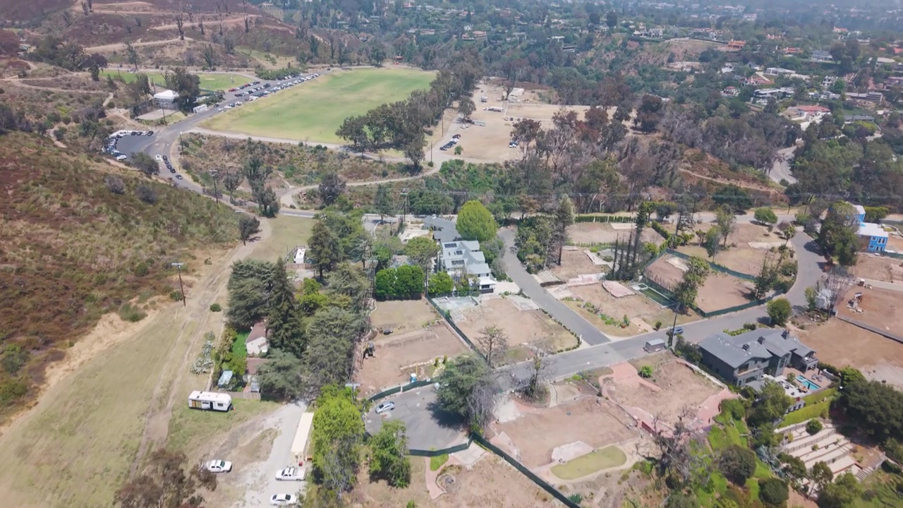 Zona suburbana con secuelas de incendio forestal mostrando árboles quemados, lotes vacíos, estructuras dispersas que sobrevivieron, un campo deportivo verde y vegetación seca en la ladera. California, EE. UU. Toma aérea amplia con inclinación ascendente.