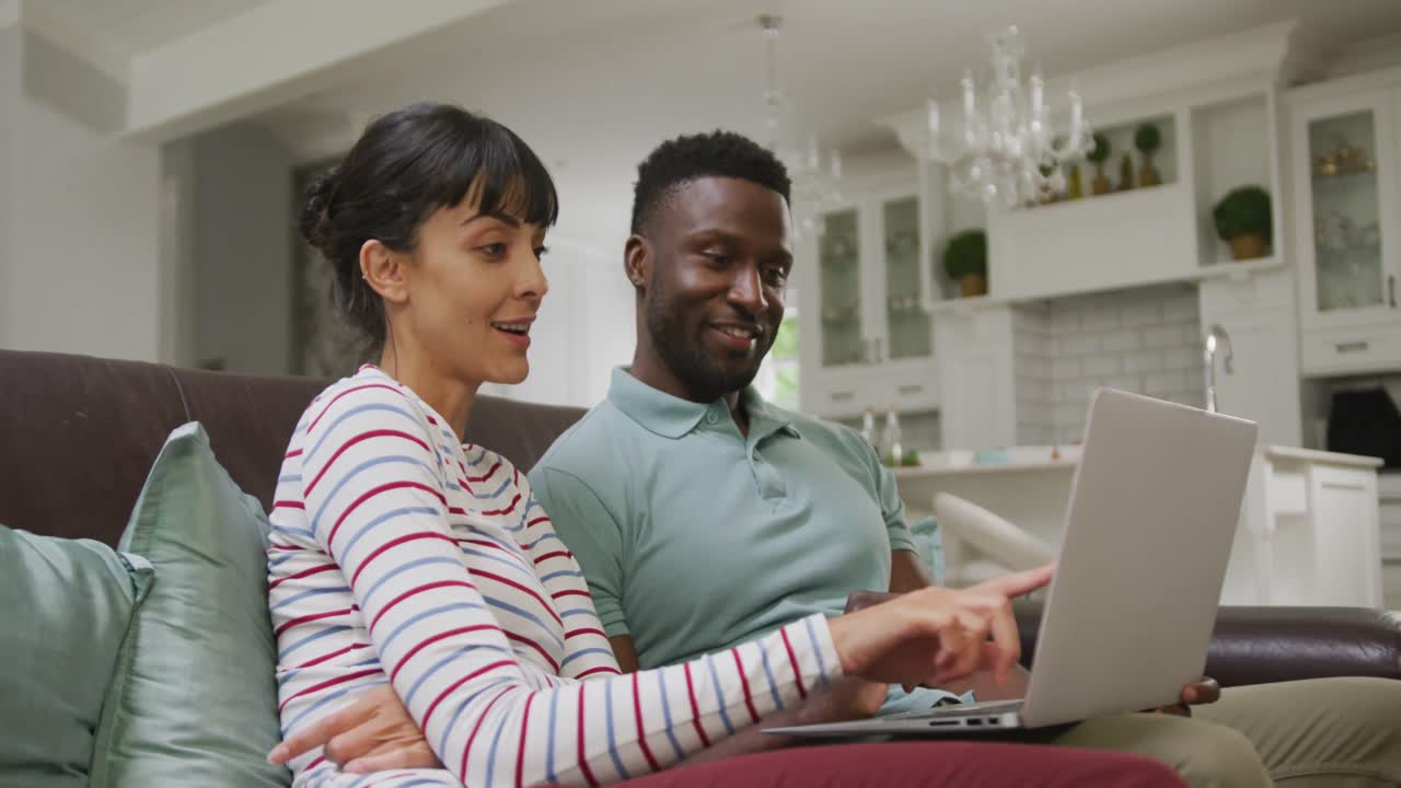 Happy diverse couple sitting on couch and using laptop in living room