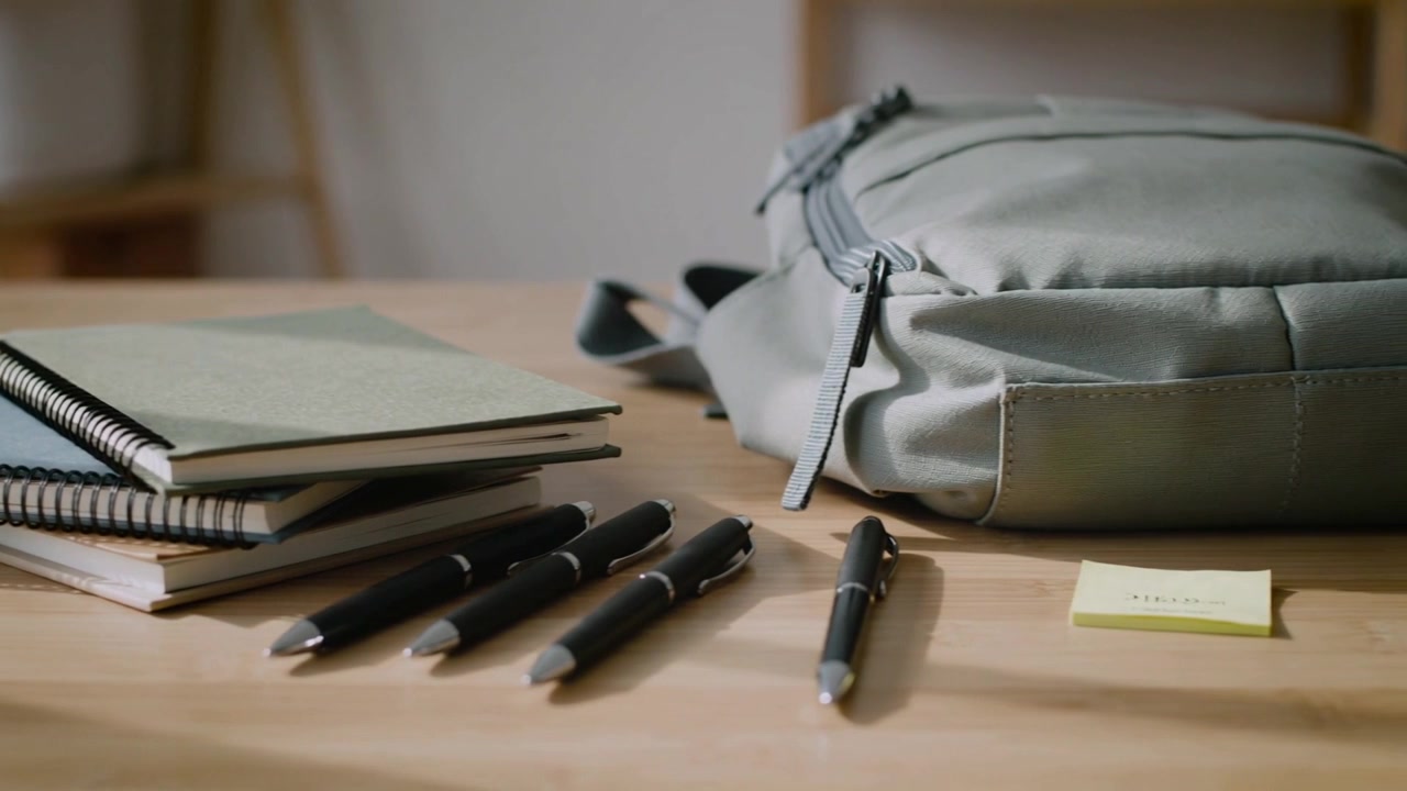 Kamera se posouvá doprava po studijním stole, odhaluje batoh, sešity, pera a lepící lístek, aby ukázala stůl.