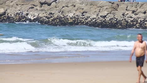 male-tourist-is-strolling-along-a-caparica-beach-with-sun-and-blue-sea,-capturing-the-beauty-of-the-scenery-with-his-camera