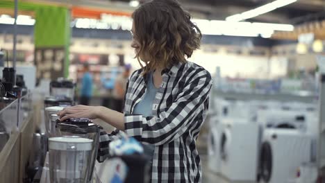 In-the-appliances-store,-a-brunette-curly-woman-in-a-plaid-shirt-chooses-a-blender-for-shopping-by-viewing-and-holding-the-device-top-in-her-hands.-Side-view