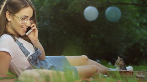 Caucasian-woman-in-glasses-lying-on-the-grass-in-the-park-and-talking-on-the-mobile-phone-near-a-kitty-cat-in-a-basket