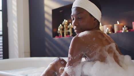 Smiling-african-american-woman-with-towel-taking-bath-and-touching-her-skin-in-bathroom