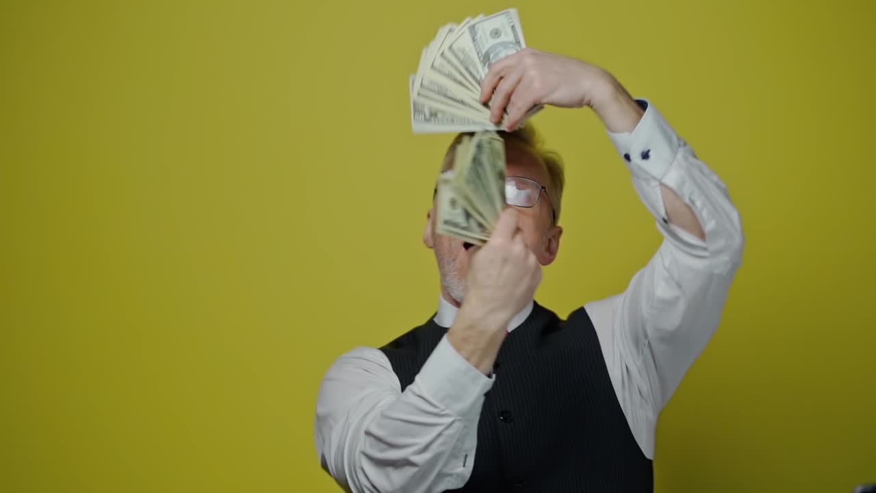 Funny senior man waving dollar banknotes. A lot of money in elderly man's hands. Happy businessman holds dollars on yellow background. Slow motion.