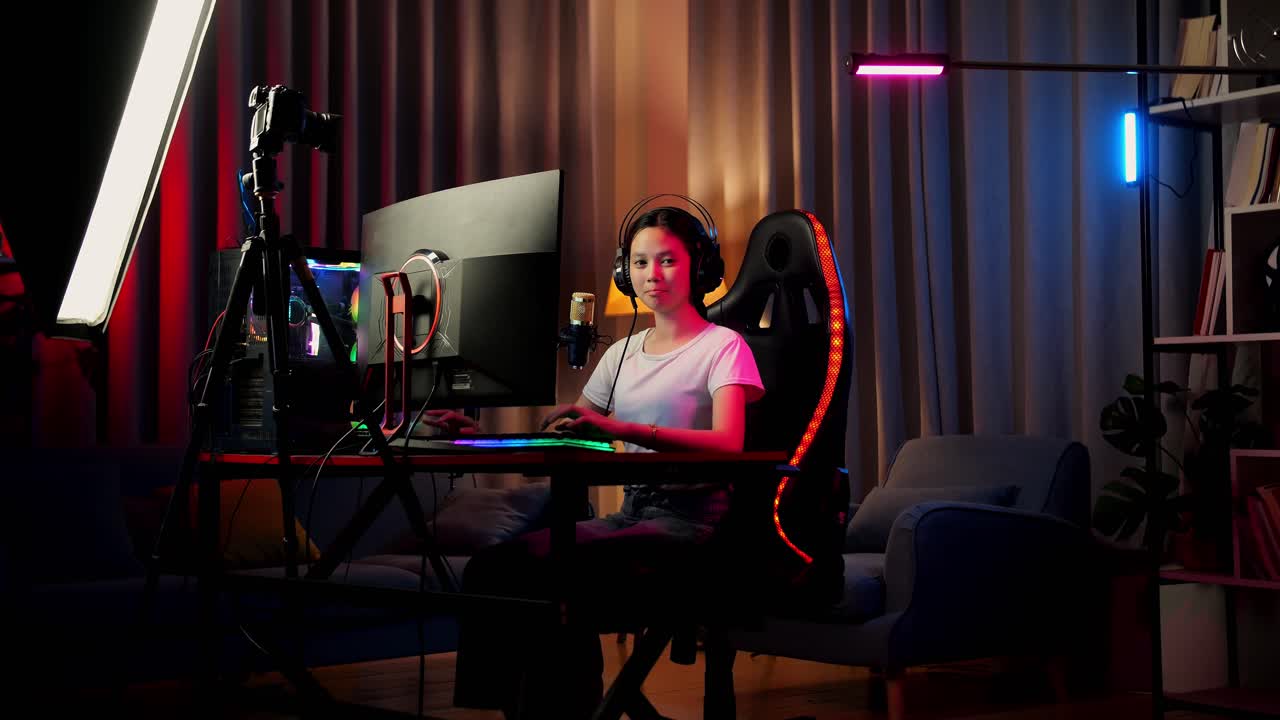 chica asiática que sonríe y cruza los brazos mientras juega un juego en red en una computadora personal. videojuego de transmisión en vivo, escritorio iluminado por luz de tira led rgb